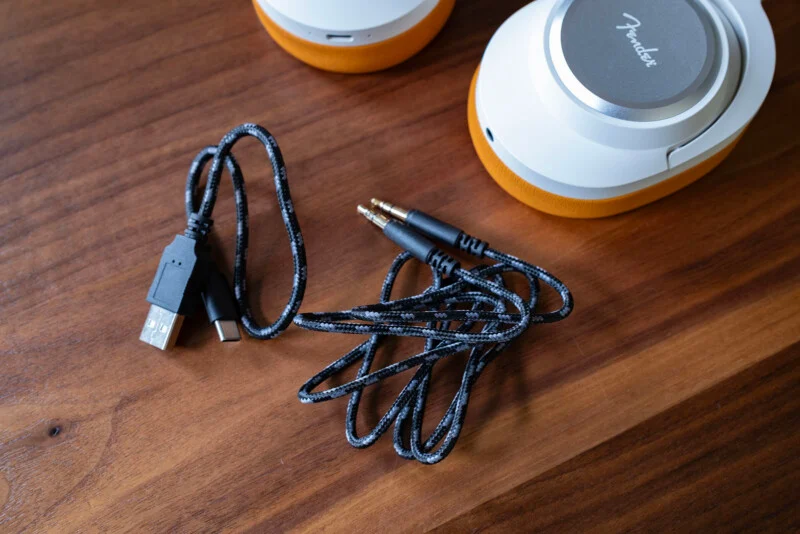 Плетений аудіокабель з двома роз'ємами 3,5 мм та чорний зарядний кабель USB-A до USB-C лежать на дерев'яній поверхні поряд з парою білих і помаранчевих навушників.