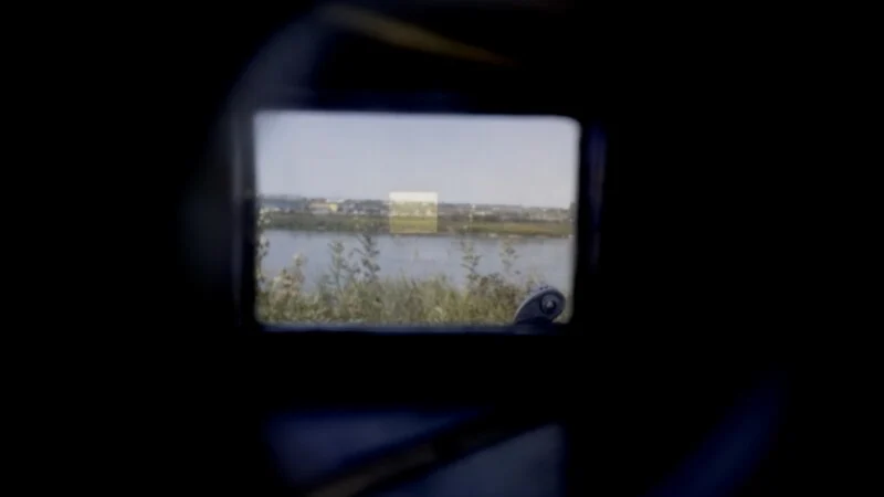 Вид через видошукач камери, що показує берег річки з високою травою, водойму та віддалені будівлі під чистим небом.
