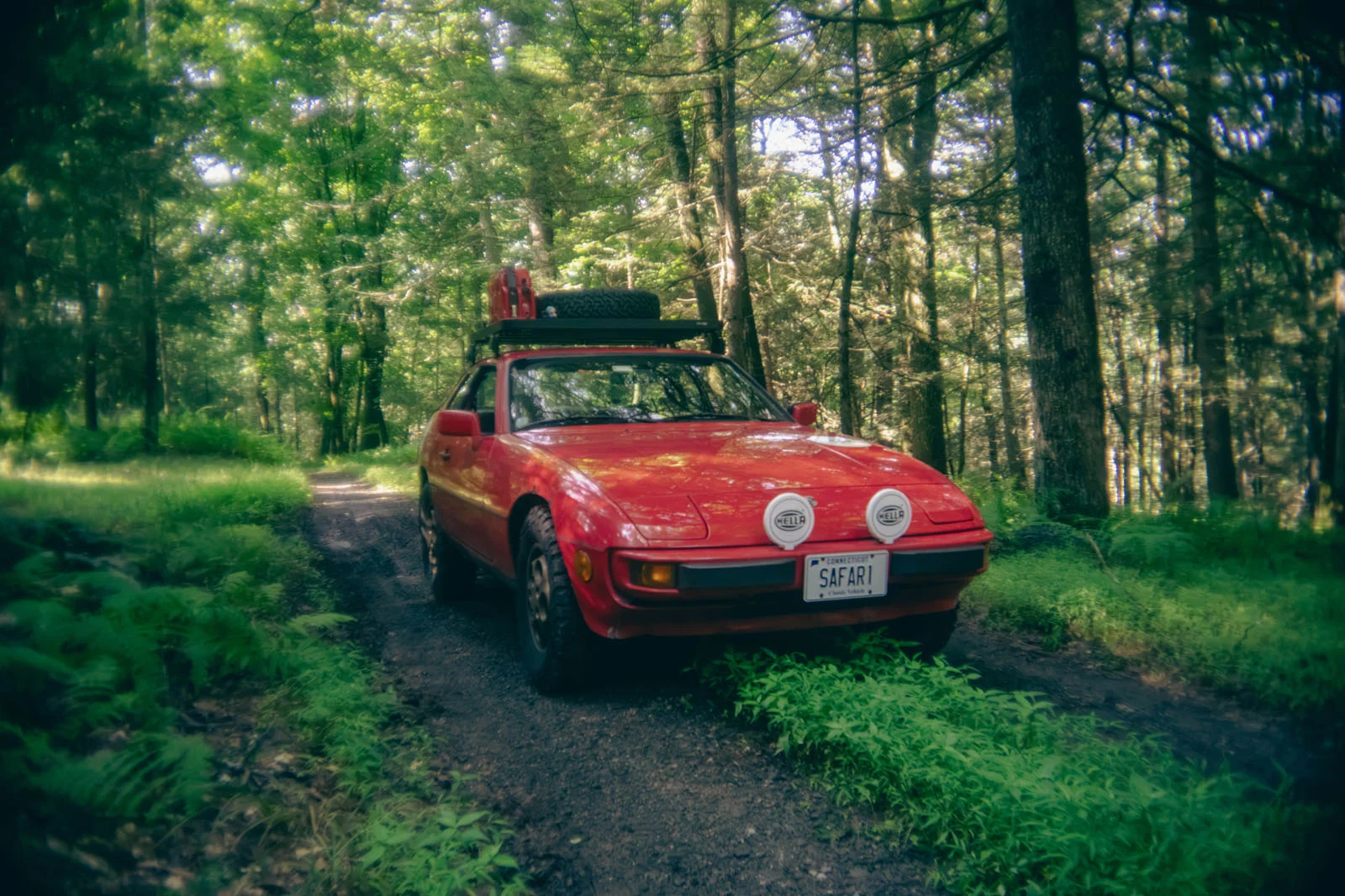 Червоний Porsche 924, модифікований для бездоріжжя з багажником на даху, запасною шиною та протитуманними фарами, стоїть на ґрунтовій дорозі в лісі.