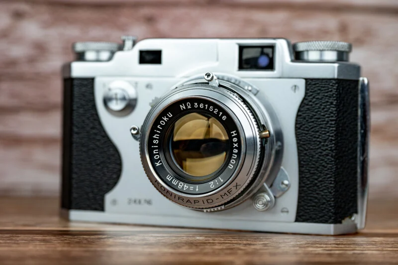 Вінтажна сріблясто-чорна плівкова камера Konishiroku Hexanon 48mm f/2 лежить на дерев'яній поверхні на фоні розмитої цегляної стіни.
