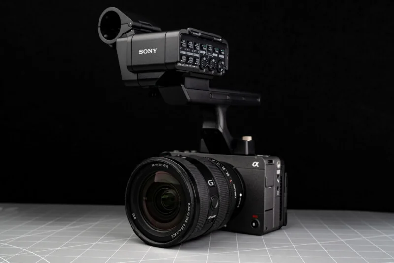 Професійна відеокамера Sony з великим об'єктивом та зовнішнім аудіоінтерфейсом розміщена на сірій поверхні з сітчастим візерунком на чорному тлі.