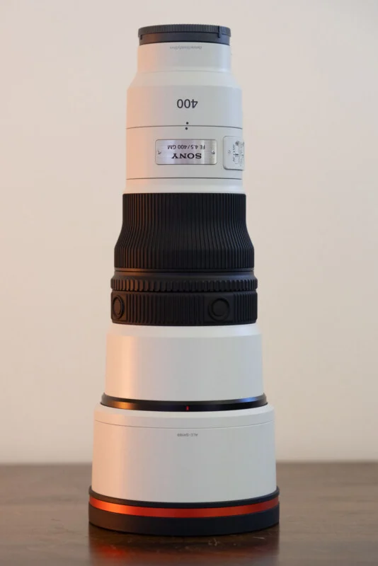 Білий телеоб'єктив Sony FE 4.5/400 GM стоїть вертикально на дерев'яній поверхні на простому тлі.