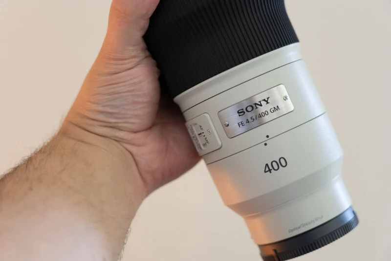 Рука людини тримає білий об'єктив Sony FE 4.5/400 GM на простому тлі.