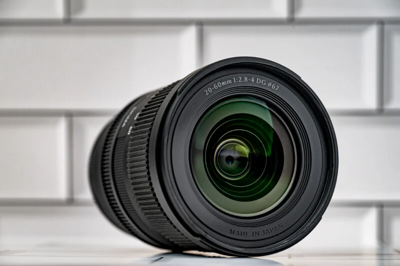 Чорний об'єктив камери зі специфікацією 20-60mm 1:2.8-4 DG 67mm лежить на білій плитці.