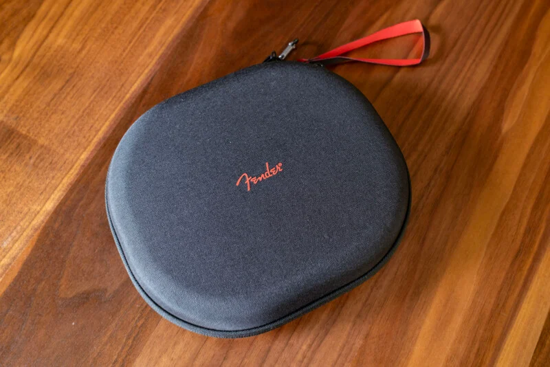 Чорний овальний чохол на блискавці з червоним логотипом "Fender" та червоним ремінцем лежить на дерев'яній поверхні.