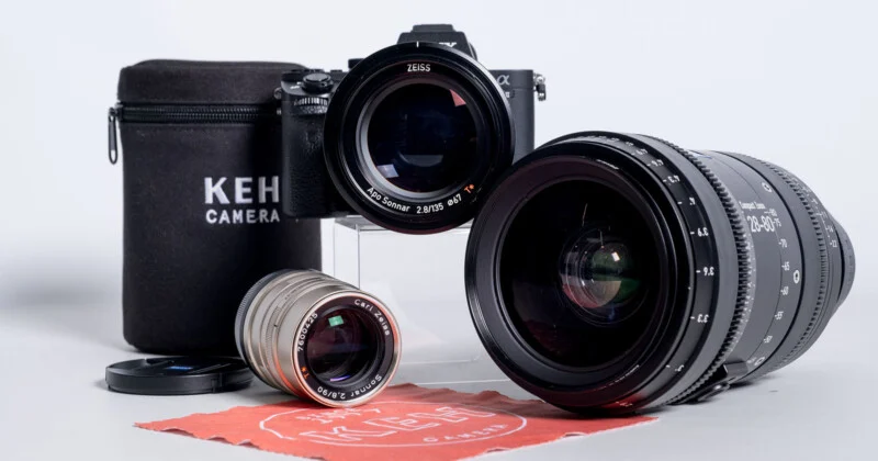 Фото камери з об'єктивом Zeiss, яка лежить поруч з меншим сріблястим об'єктивом, великим чорним зум-об'єктивом, чохлом з логотипом KEH Camera та червоною серветкою для чищення.