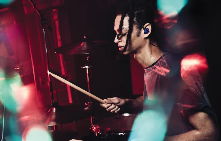 A drummer wearing a pair of blue Sony IER-M500 IEMs while sweatily performing on stage under colored lights