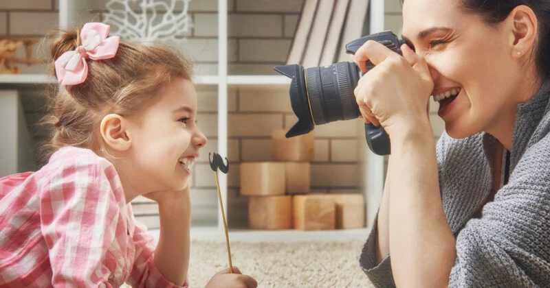 Жінка посміхається, фотографуючи маленьку дівчинку, яка тримає вуса на паличці.