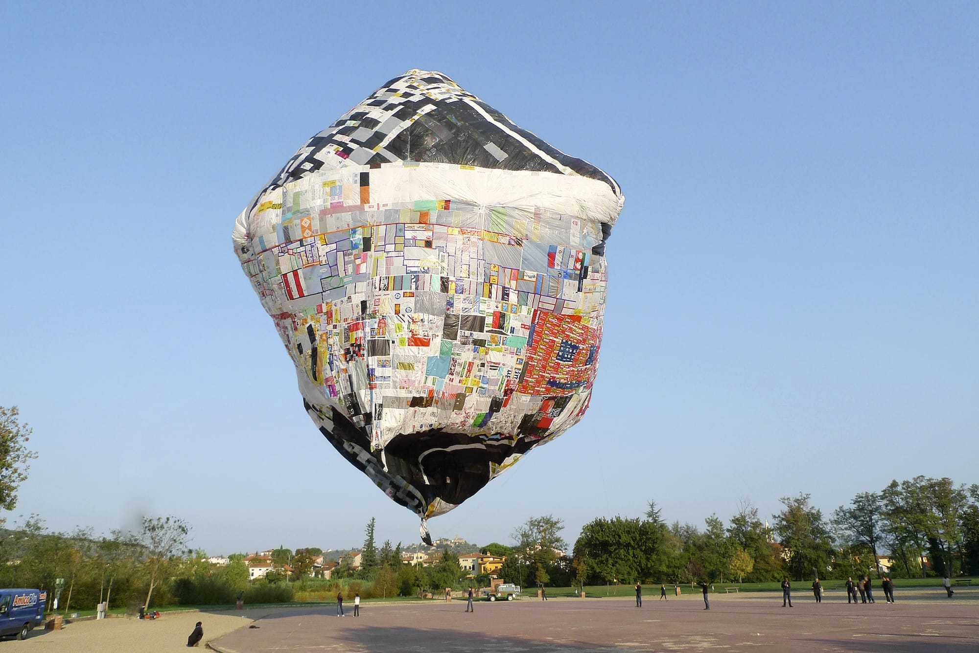 a floating artwork by Tomás Saraceno featuring a patchwork object resembling an unusual air balloon over a square