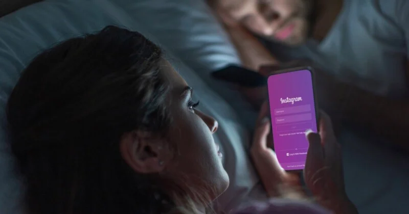 Жінка лежить у ліжку вночі, дивлячись на свій смартфон, на якому відображається екран входу в Instagram, а на задньому плані лежить чоловік.