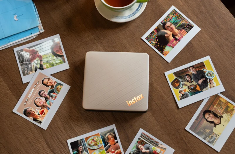 Бежевий принтер Instax стоїть на дерев'яному столі, оточений кількома миттєвими фотографіями, картою та чашкою чаю.