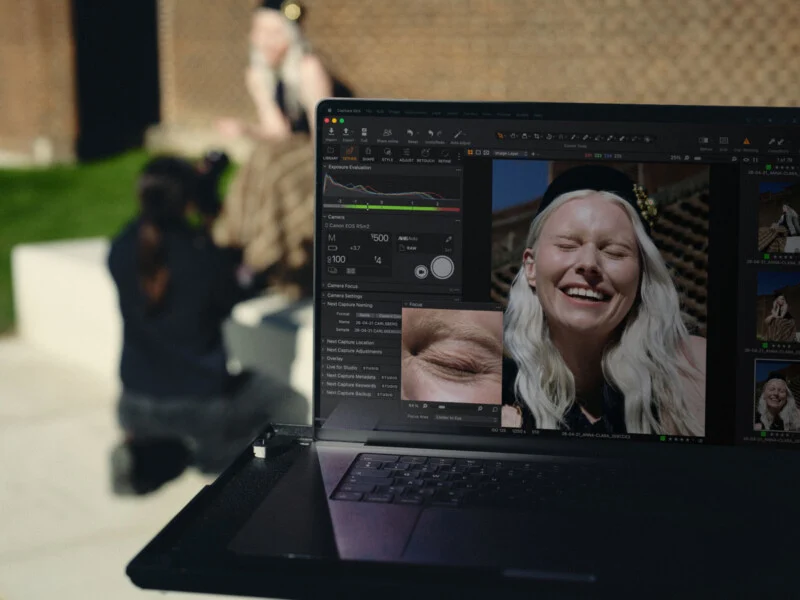 Екран ноутбука з програмним забезпеченням Capture One, що показує портрет усміхненої жінки. На розмитому фоні видно фотографа та модель.