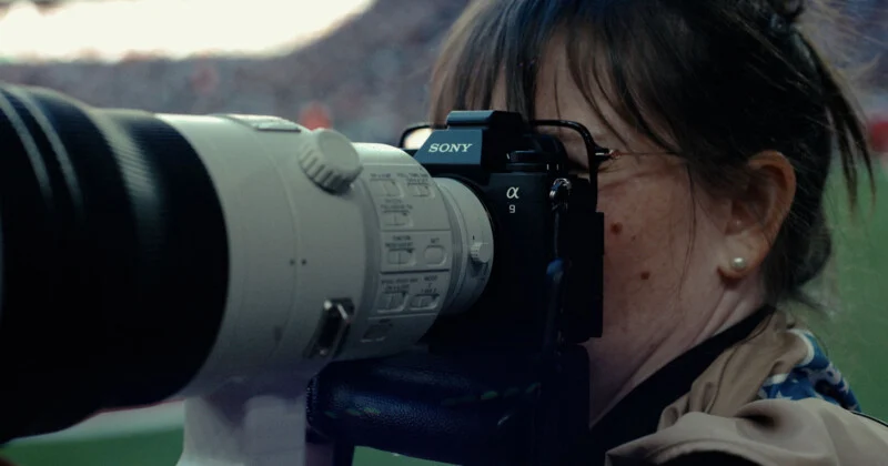 Людина з темним волоссям та в окулярах дивиться у видошукач камери Sony Alpha 9 з великим телеоб’єктивом.
