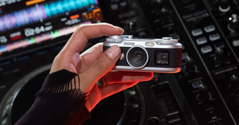 Рука в чорному рукаві тримає сріблясто-чорну камеру Instax перед мікшерною консоллю DJ.