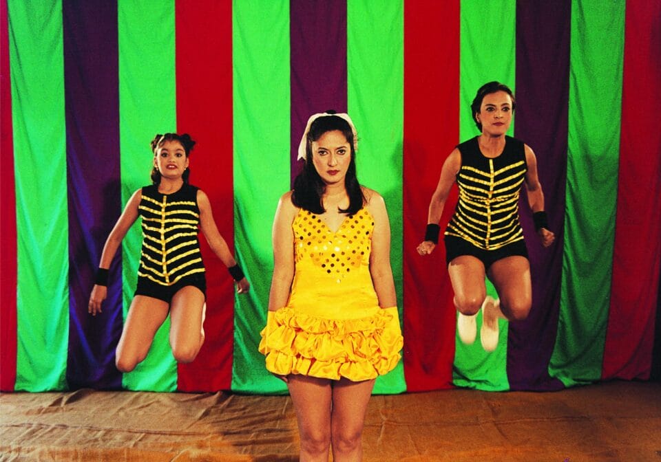 a phtoograph by Pushpamala N. of three wimen in gold and black costumes against a striped background. two women on either side of the subject in the middle are mid-jump