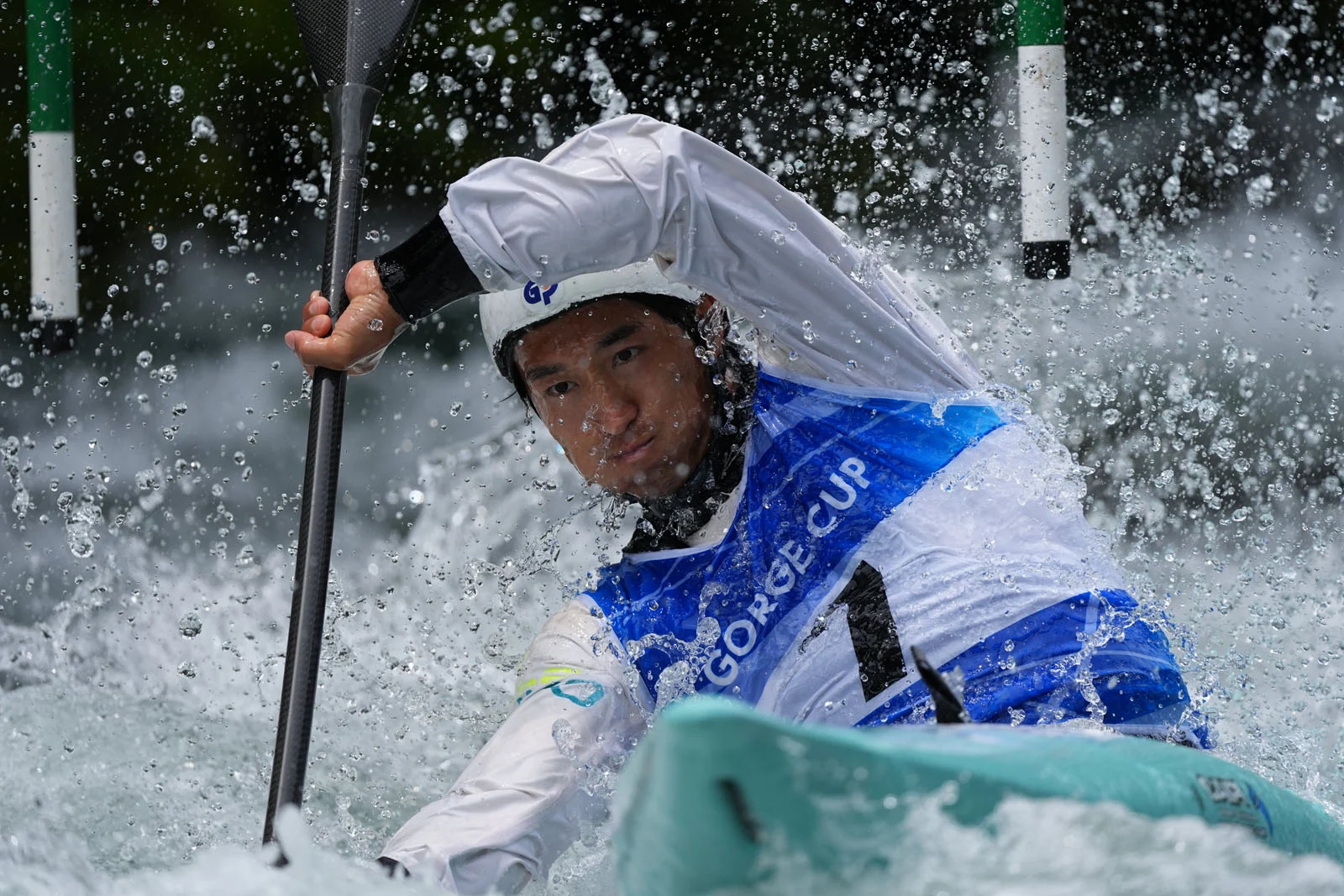 Каякер у білій та синій футболці з написом "GORGE CUP" пливе крізь бурхливу воду між двома слаломними жердинами.