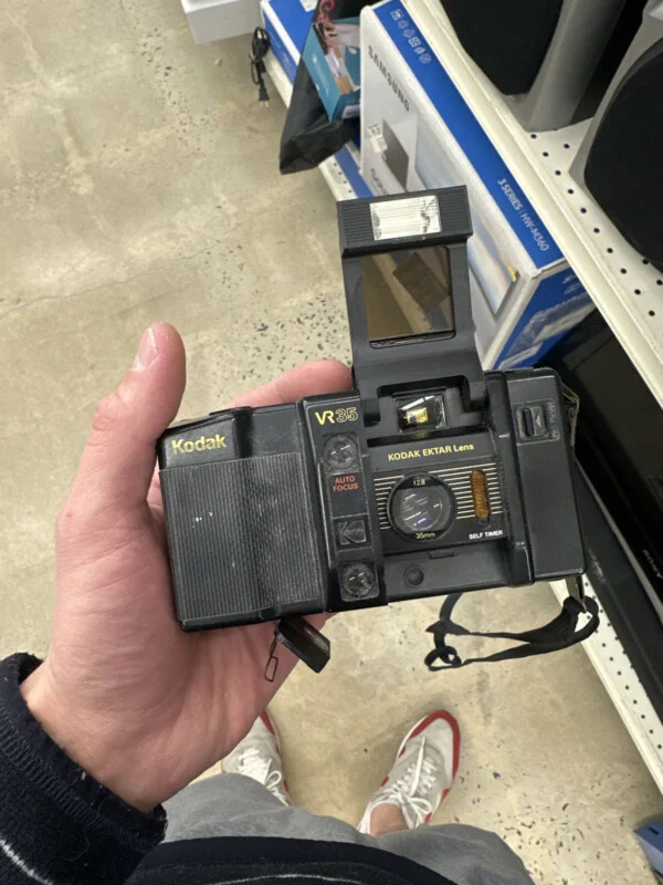 Рука тримає чорну плівкову камеру Kodak VR35 з піднятим спалахом в проході магазину.