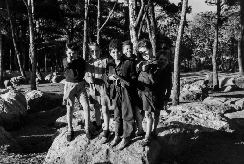 Шість хлопчиків стоять на великому камені в лісистій місцевості, позуючи з іграшковими рушницями, спрямованими в камеру.