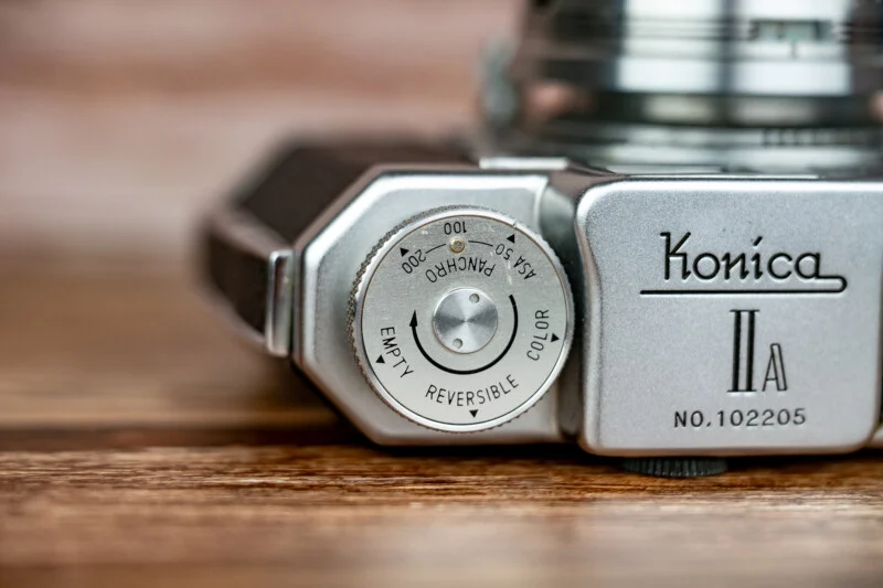 Великий план верхньої панелі сріблястої камери Konica IIA, що показує диск нагадування про плівку з позначками ISO та типами плівки, розташований на дерев'яній поверхні.