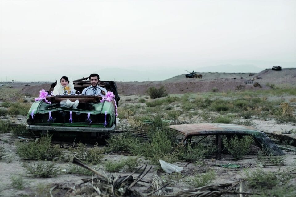 a photograph by Gohar Dashti of a newlywed couple sitting in a car in a junkyard. a military tank looms in distance