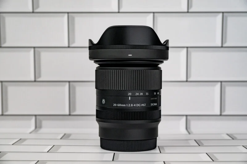 Чорний об'єктив Sigma 20-60mm стоїть вертикально на фоні білої плитки.