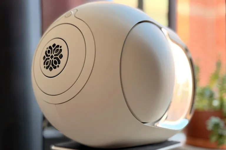 A Light Pearl Devialet Phantom Ultimate 108 dB wireless speaker sitting on a stand in front of a window