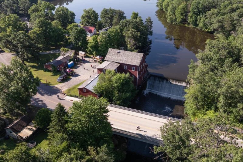 Аерофотозйомка показує червону будівлю млина поруч із водоспадом та міст, де велосипедисти переїжджають річку.