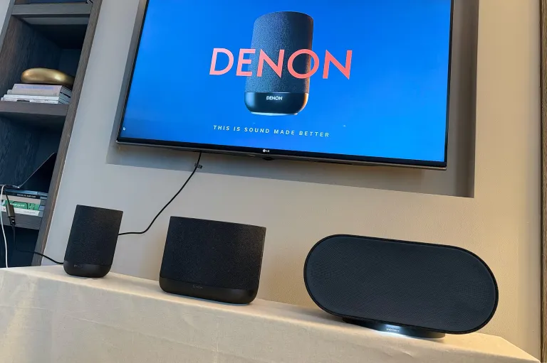 The DENON Home 200, 400, and 600 Dolby Atmos-compatible wireless connected whole-home speaker collection on a table in front of a TV showing a DENON promotional loop