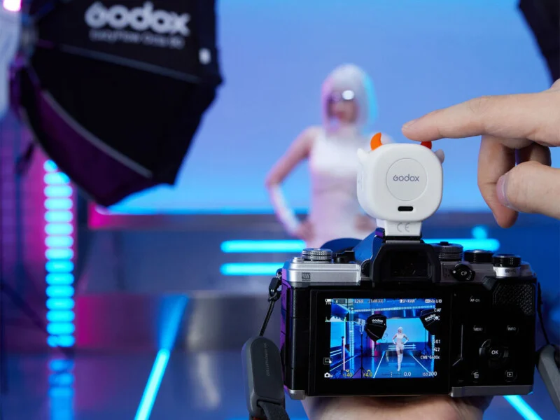Рука натискає на маленький білий бездротовий передавач Godox з ріжками диявола, встановлений зверху на цифровий фотоапарат, на якому відображається живе зображення моделі в студії.