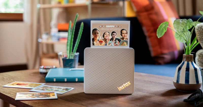 Білий принтер Instax стоїть на дерев'яному столі. У слоті видніється надрукована фотографія з написом "Happy Anniversary". Навколо розкладені інші фотографії, маленький кактус і горщикова рослина.