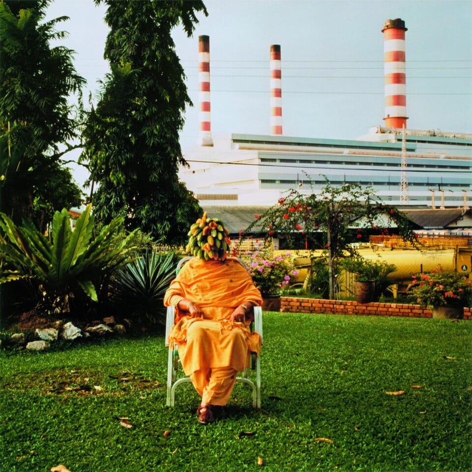 a vibrant photo by Simryn Gill of a woman sitting on a plastic chair in a lawn. her face is covered with a bunch of gourds