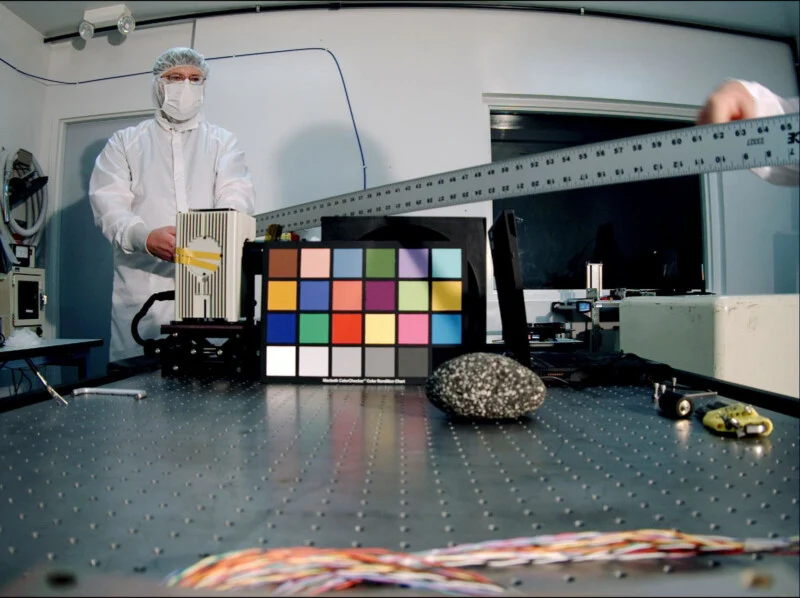 Людина в білому костюмі чистої кімнати стоїть за столом, тримаючи металеву лінійку, на поверхні якої лежить кольорова калібровочна діаграма та строкатий камінь.