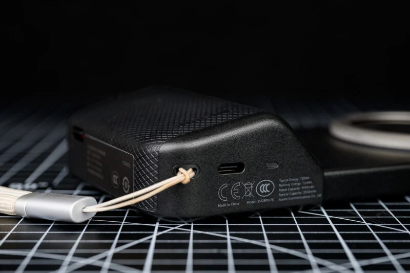 Макро знімок чорного електронного пристрою з текстурованою поверхнею, портом USB-C, вигравіруваними написами та прикріпленим ремінцем, розташований на поверхні з сітчастою основою, на розмитому тлі.