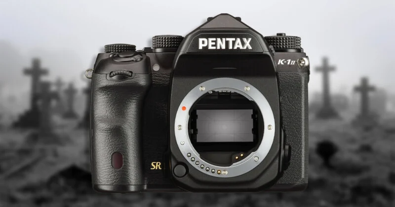 Чорна дзеркальна камера Pentax K-1 Mark II на розмитому чорно-білому фоні кладовища з хрестами.