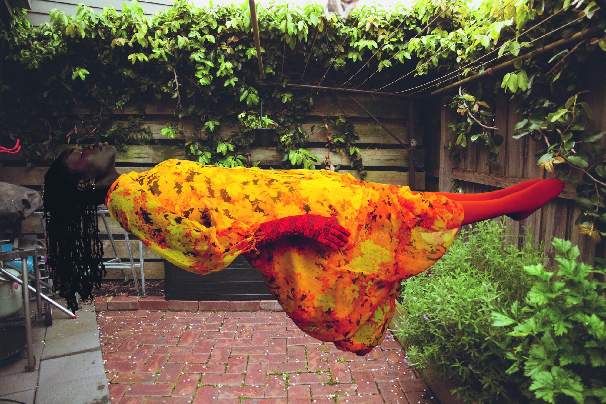 a photograph by Atong Atem of a woman wearing a warm-hued floral dress levitating as she lays horizontally