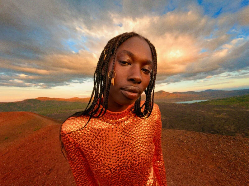 Людина з плетеним волоссям та золотими аксесуарами для волосся одягнена в мерехтливий помаранчевий топ, розшитий блискітками, стоячи на скелястому, червонуватому ландшафті на заході сонця.