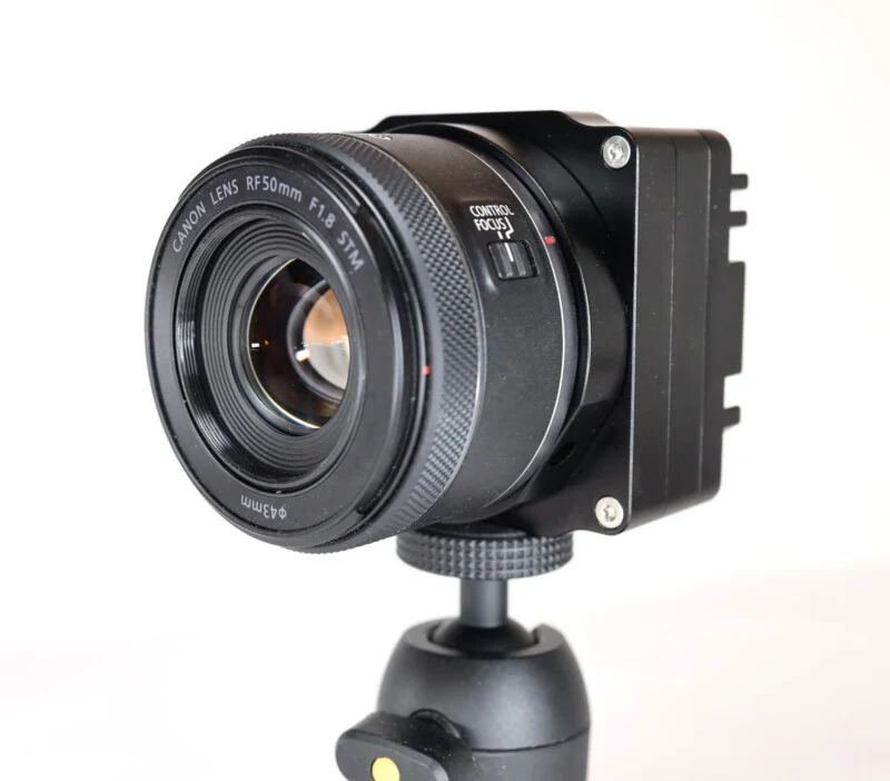 Об'єктив Canon RF 50mm F1.8 STM, приєднаний до чорного корпусу камери прямокутної форми, встановленої на невеликій штативній головці на білому тлі.