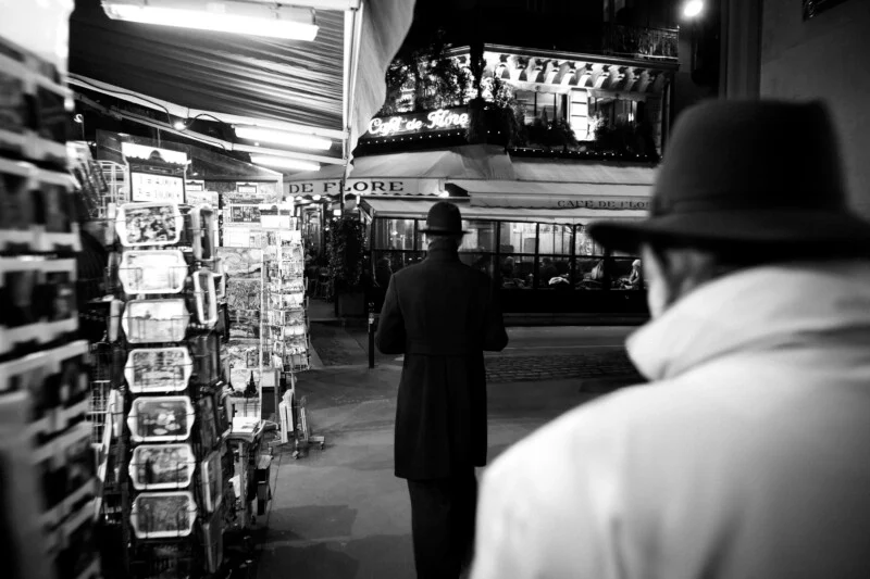 Людина в капелюсі та пальті йде до Café de Flore вночі, видно з-за спини іншої людини на передньому плані.