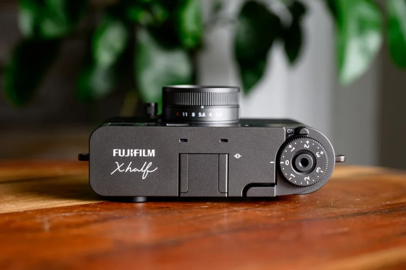 Вигляд зверху чорної камери Fujifilm X-H2, що лежить на дерев'яній поверхні, показуючи об'єктив, регулювальні кільця та бренд Fujifilm, з розмитими зеленими листям на задньому плані.
