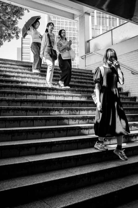 Чотири жінки йдуть по зовнішніх сходах в міській місцевості; троє спускаються, одна тримає парасольку, а інша жінка в сукні піднімається, освітлена сонцем. Сцена в чорно-білому кольорі.
