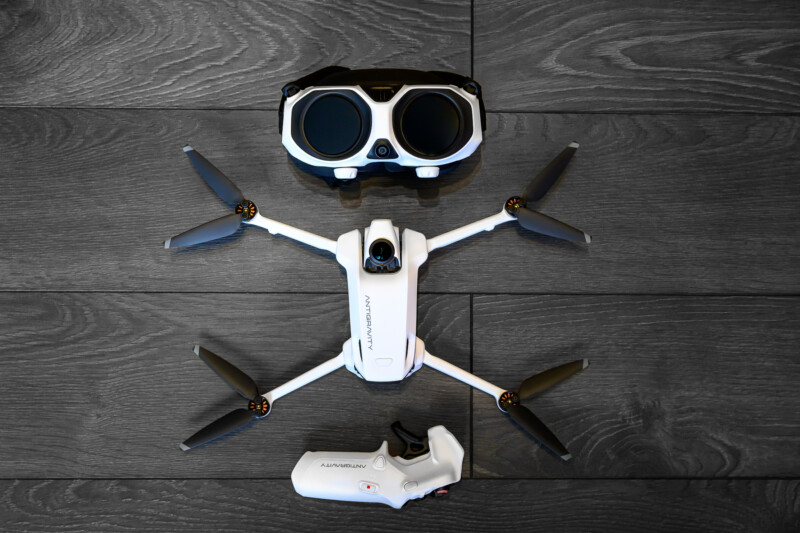 Білий дрон, білий контролер і пара окулярів FPV чорно-білого кольору акуратно розставлені на темній дерев'яній підлозі. Дрон знаходиться посередині, окуляри – вище, а контролер – нижче.