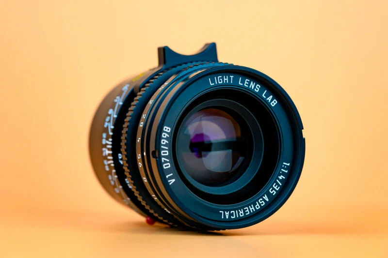 Крупний план фотооб'єктива з написом "Light Lens Lab 1:1.2/35 ASPH EIRCIAL VI 070198" на гладкому персиковому фоні. Об'єктив розташований вертикально, обличчям до глядача.