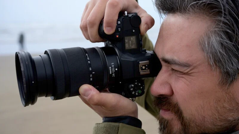 Людина з сивіючим волоссям робить фотографію за допомогою дзеркальної камери з великим об'єктивом, тримаючи її біля ока. Людина зосереджена, на задньому плані розмитий пляж та інша людина.