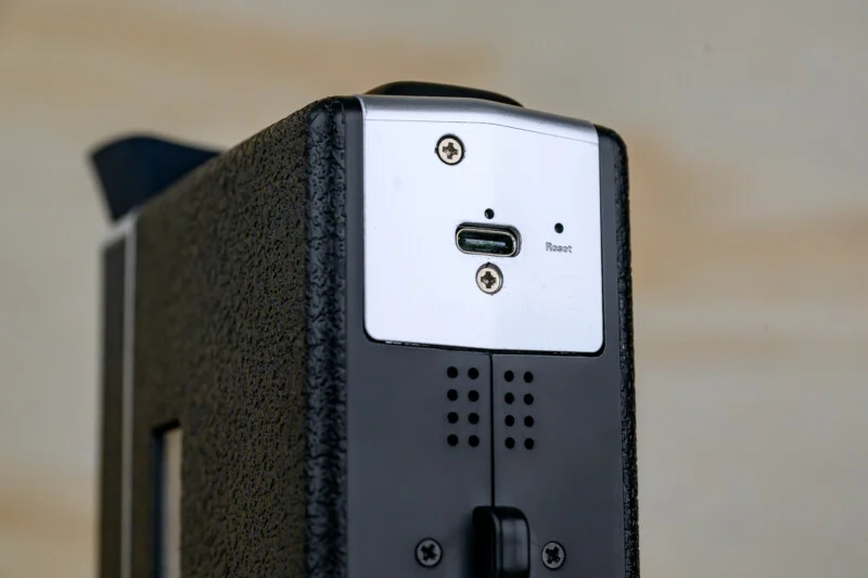 Крупний план чорно-сріблястого електронного пристрою, що показує порт USB-C, маленьку кнопку скидання та два гвинти на текстурованій поверхні.