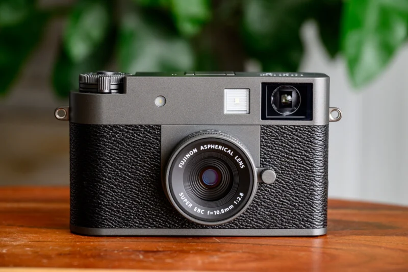 Чорна камера Fujinon Rangefinder з текстурованим хватом лежить на дерев'яній поверхні, на задньому плані розмиті зелені листя.