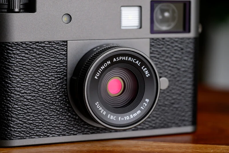 Великим планом об'єктив камери, що показує Fujinon Aspherical Lens, Super EBC f=10.8mm 1:2.8, встановлений на текстурованому чорно-сріблястому корпусі камери, з видимими частинами видошукача та спалаху.