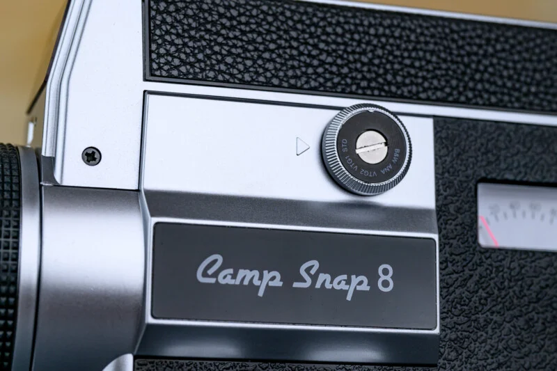Крупний план вінтажної камери з написом "Camp Snap 8", що показує чорно-сріблястий корпус з диском і частиною датчика.