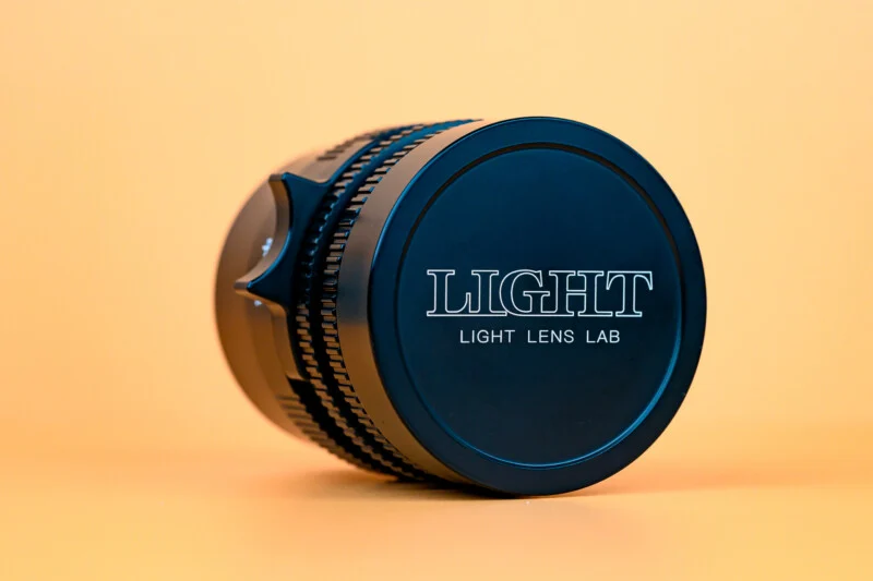 Чорний фотооб'єктив з написом "LIGHT LIGHT LENS LAB" на передній кришці, покладений на бік на тлі суцільного світло-помаранчевого фону.