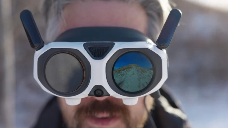 Крупний план людини у великих білих окулярах VR або FPV з антенами. Відбиття в одній лінзі показує засніжений гірський пейзаж під блакитним небом. Фон розмитий.