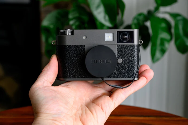 Людина тримає камеру Fujifilm з ковпачком на об'єктиві, на задньому плані розмиті зелені рослини та біла стіна.